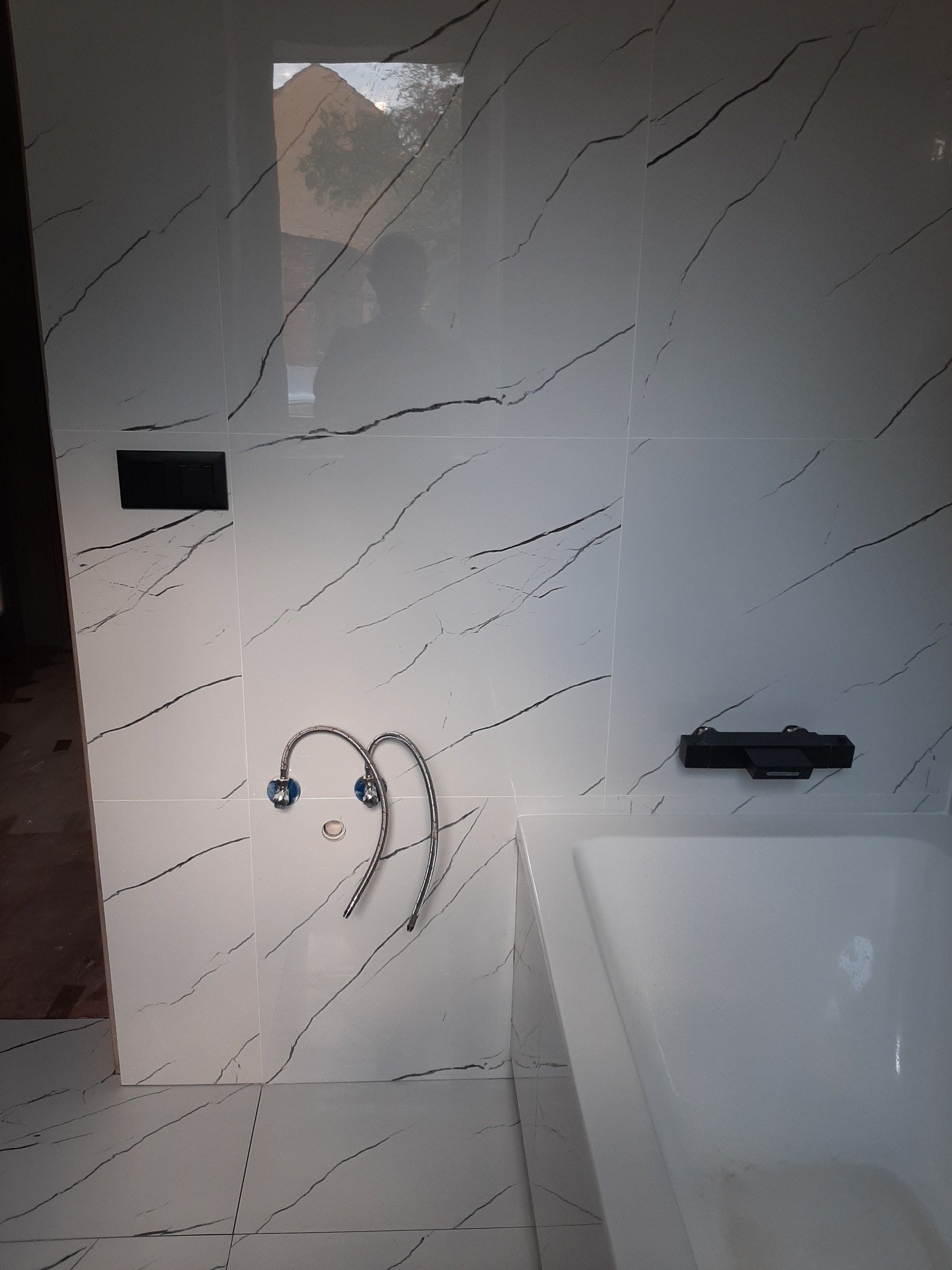 Biała łazienka z płytkami imitującymi marmur, chromowana bateria umywalkowa z niebieskimi akcentami, widoczna wanna i czarny panel sterowania.