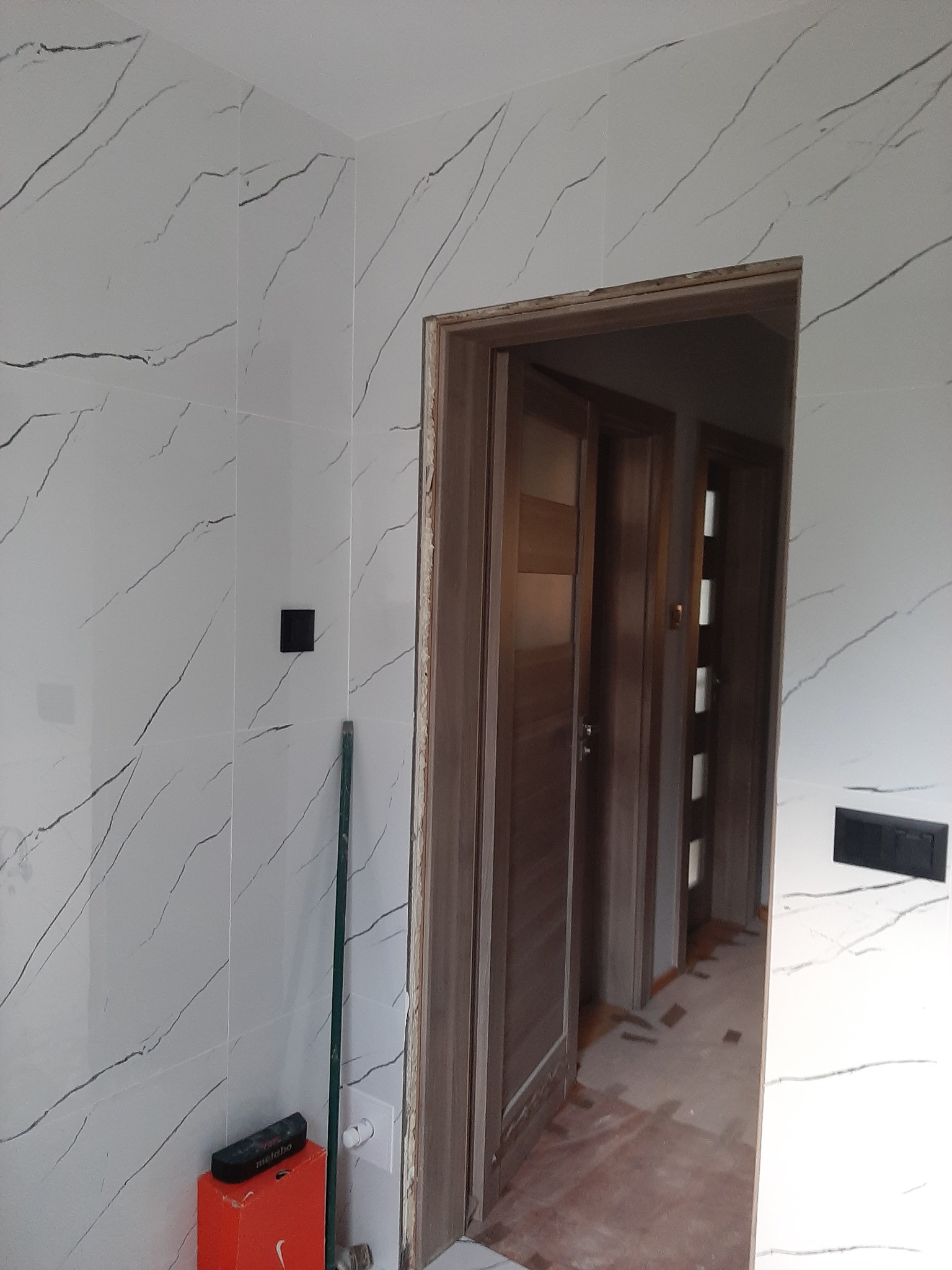 Ściana wyłożona białymi płytkami imitującymi marmur z czarnymi żyłkami, widoczny fragment ościeżnicy drzwiowej oraz podłoga w trakcie prac wykończeniowych, w rogu ściany narzędzia i pudełko po butach.