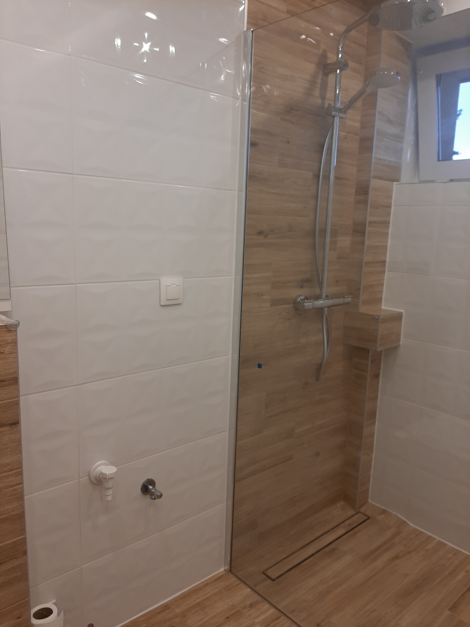 Nowoczesna kabina prysznicowa walk-in z odpływem liniowym, wykończona białymi płytkami strukturalnymi i panelami imitującymi drewno, wyposażona w deszczownicę i słuchawkę prysznicową.