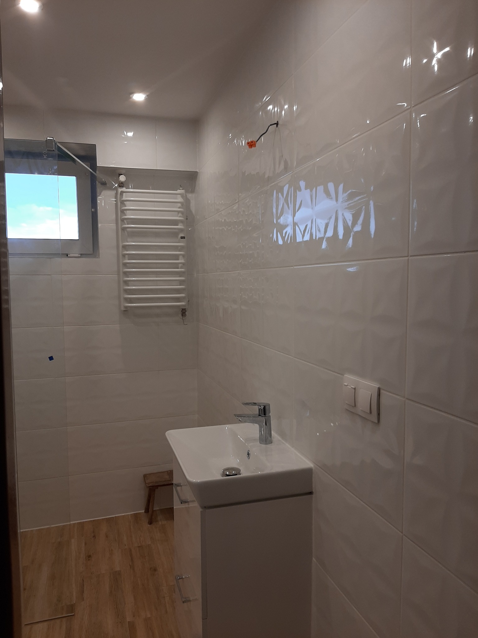 Biała łazienka z umywalką na szafce, grzejnikiem drabinkowym i błyszczącymi, wzorzystymi płytkami na ścianach, drewnopodobna podłoga, mały stołek w rogu.