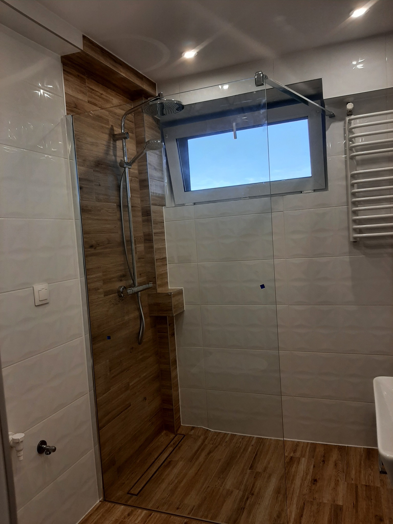 Nowoczesna kabina prysznicowa z drewnopodobnymi płytkami, szklaną ścianką i oknem z widokiem na błękitne niebo.
