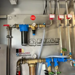 System filtracji i rur z manometrami zamontowany na metalowej ścianie, widoczne filtry z niebieskimi obudowami, mosiężne zawory i rury ze stali nierdzewnej, naklejka 'made by EHRLE made in Germany'...