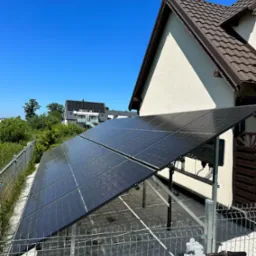 Panele fotowoltaiczne zamontowane na konstrukcji obok domu jednorodzinnego, widok zza roweru z zieloną ramą i szarymi kierownicami, w tle błękitne niebo.