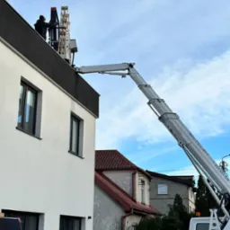 Montaż paneli fotowoltaicznych na dachu domu jednorodzinnego przy użyciu podnośnika koszowego Baltic Lift, widok z dołu na pracownika w trakcie instalacji.