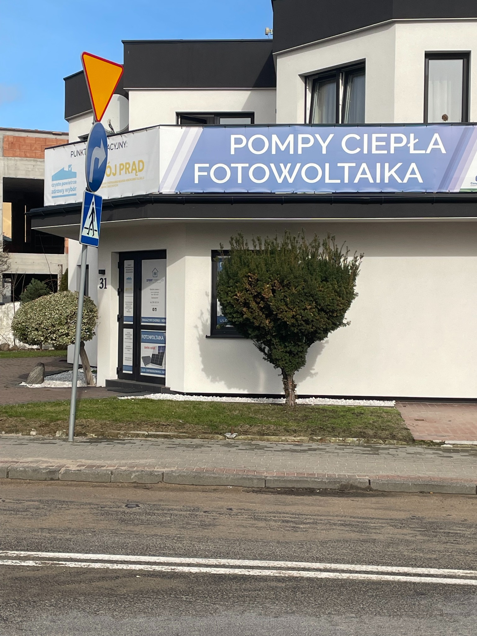 Budynek firmy z banerem reklamowym 'Pompy ciepła, Fotowoltaika', widoczny z ulicy, z numerem 31 i małym drzewkiem przed wejściem.