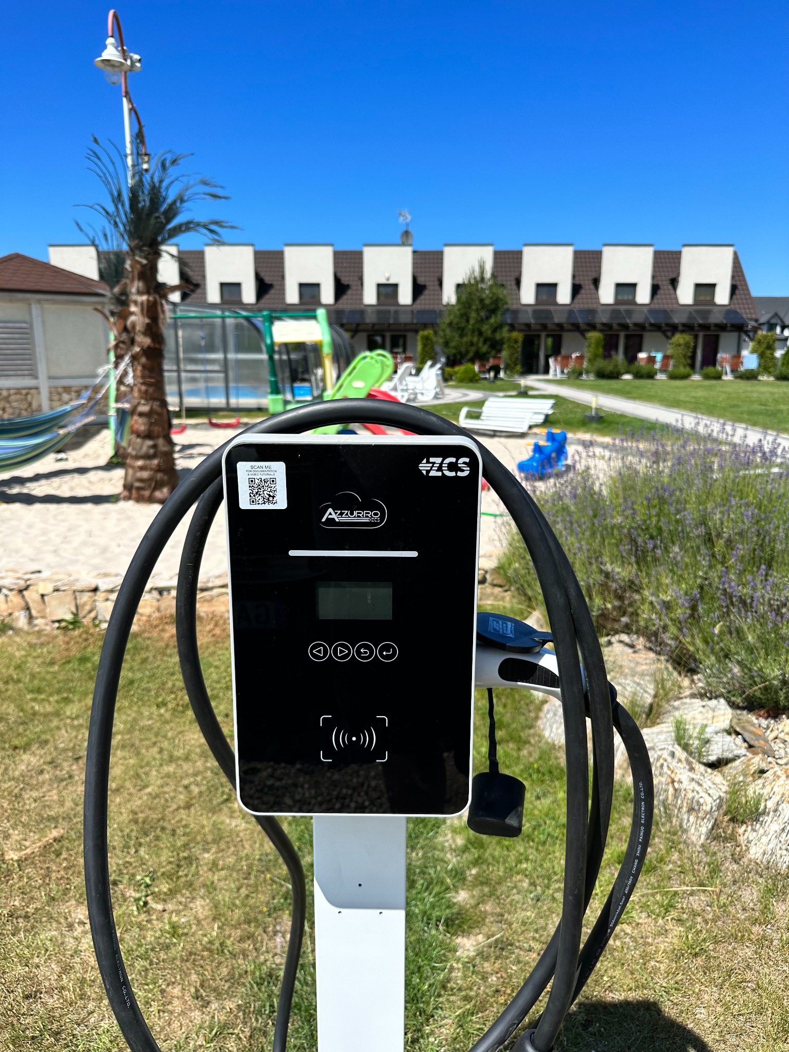 Stacja ładowania pojazdów elektrycznych Azzurro ZCS z QR kodem, na tle letniego krajobrazu z palmą i budynkiem z panelami słonecznymi.