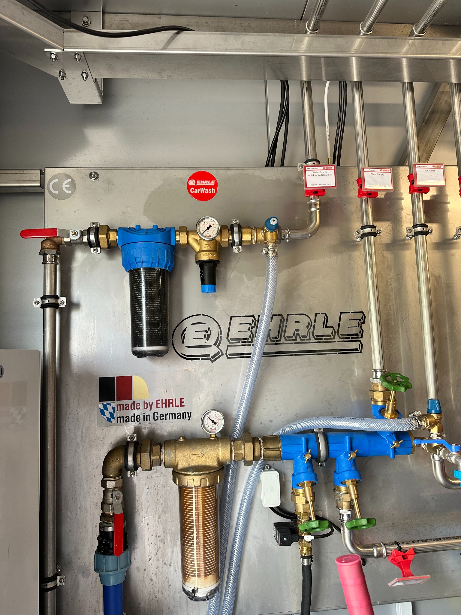 System filtracji i rur z manometrami zamontowany na metalowej ścianie, widoczne filtry z niebieskimi obudowami, mosiężne zawory i rury ze stali nierdzewnej, naklejka 'made by EHRLE made in Germany'...