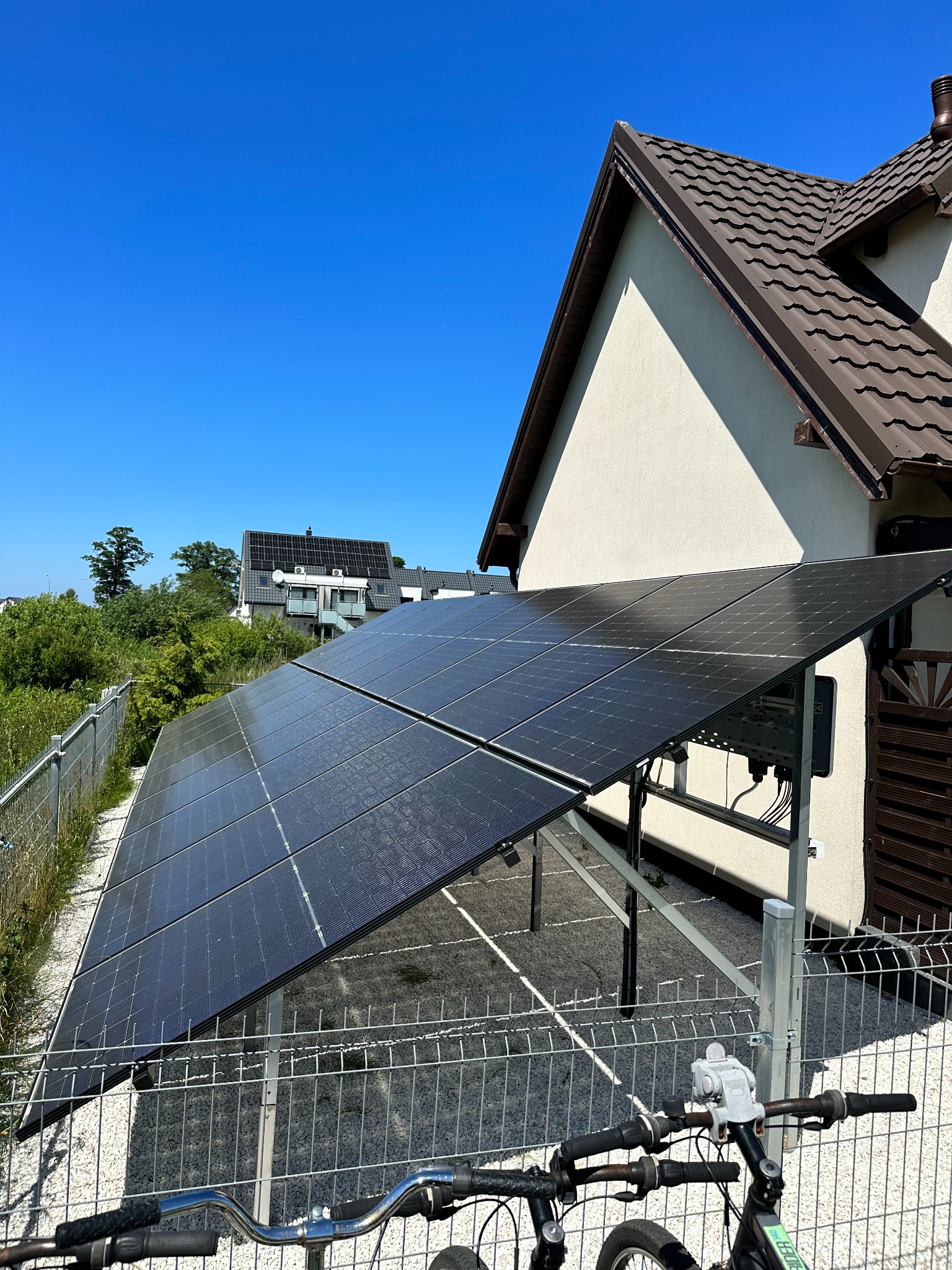 Panele fotowoltaiczne zamontowane na konstrukcji obok domu jednorodzinnego, widok zza roweru z zieloną ramą i szarymi kierownicami, w tle błękitne niebo.
