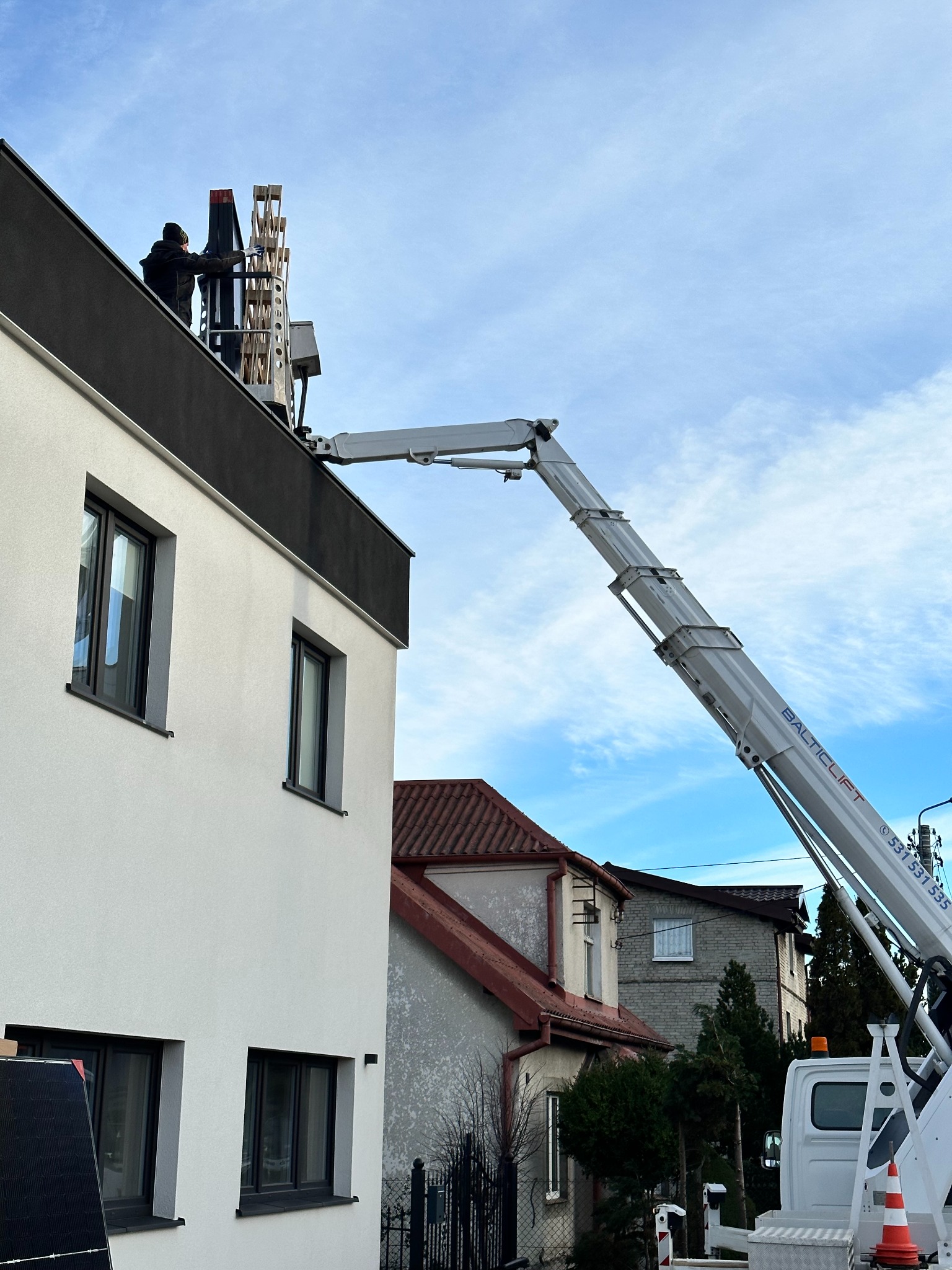 Montaż paneli fotowoltaicznych na dachu domu jednorodzinnego przy użyciu podnośnika koszowego Baltic Lift, widok z dołu na pracownika w trakcie instalacji.