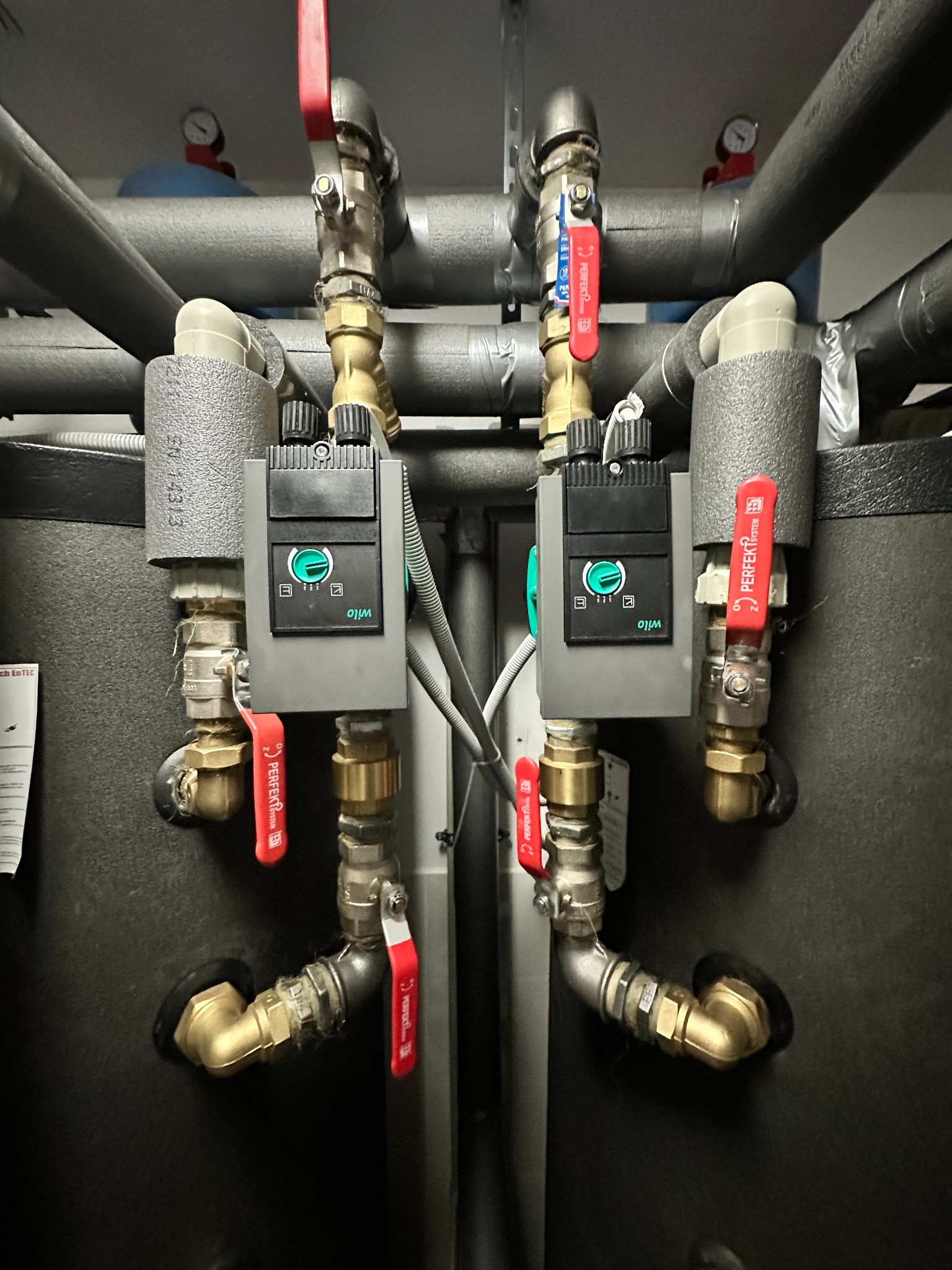 Dwa czarne zbiorniki z izolacją termiczną i pompami obiegowymi Wilo, połączone rurami miedzianymi i zaworami kulowymi z czerwonymi uchwytami, widoczne manometry na rurach.