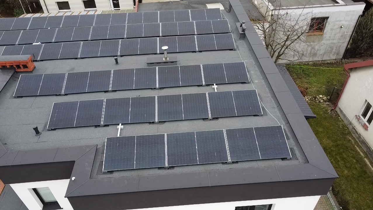 Widok z góry na instalację paneli fotowoltaicznych na płaskim dachu budynku z szarą papą, kominem i elementami wentylacyjnymi. W tle budynki mieszkalne i drzewa bez liści.