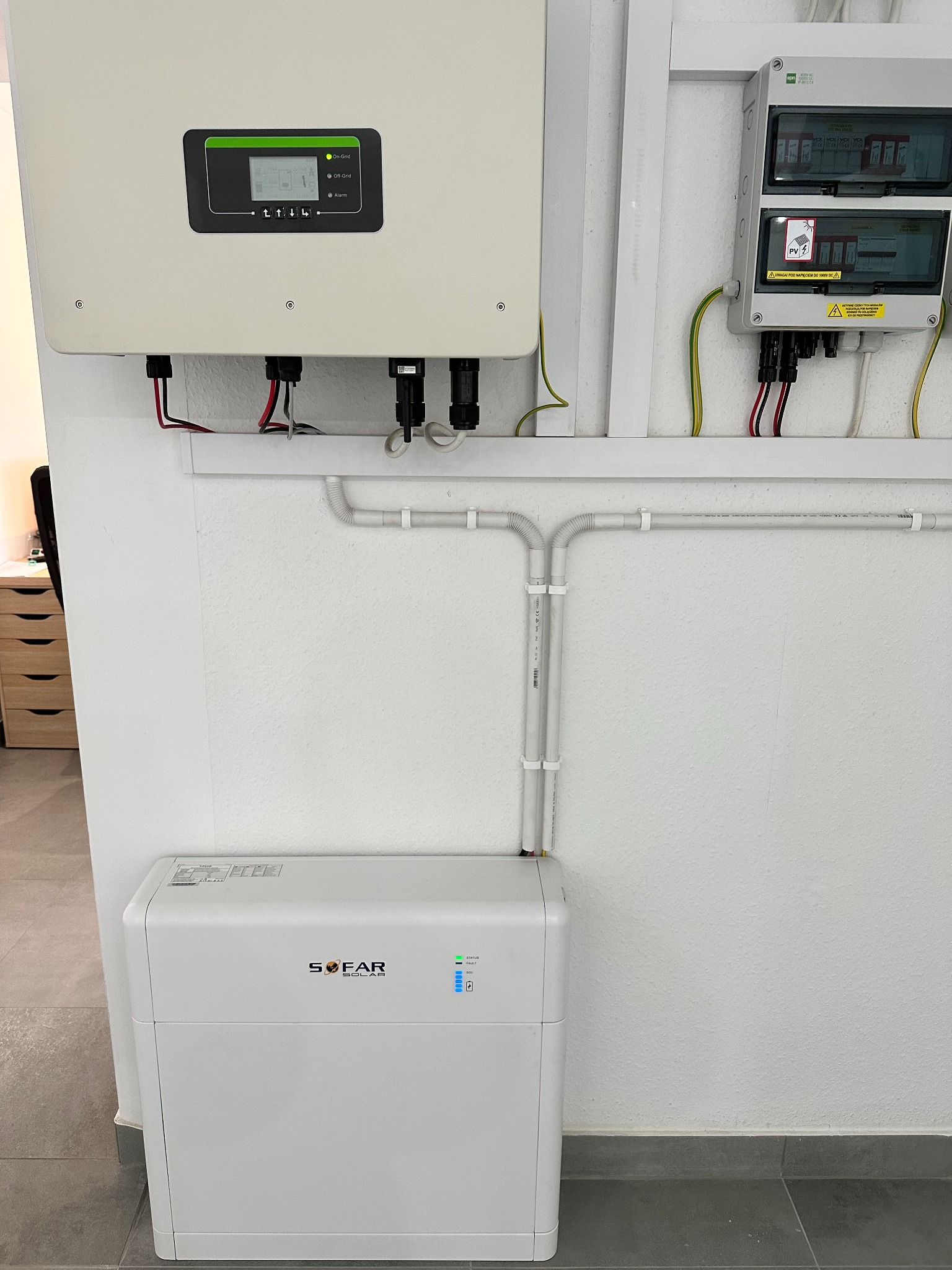 Biały falownik i akumulator energii słonecznej firmy SOFAR zamontowane na białej ścianie, widoczne przewody, rury instalacyjne i skrzynka bezpiecznikowa. Na falowniku świecą się diody sygnalizujące...
