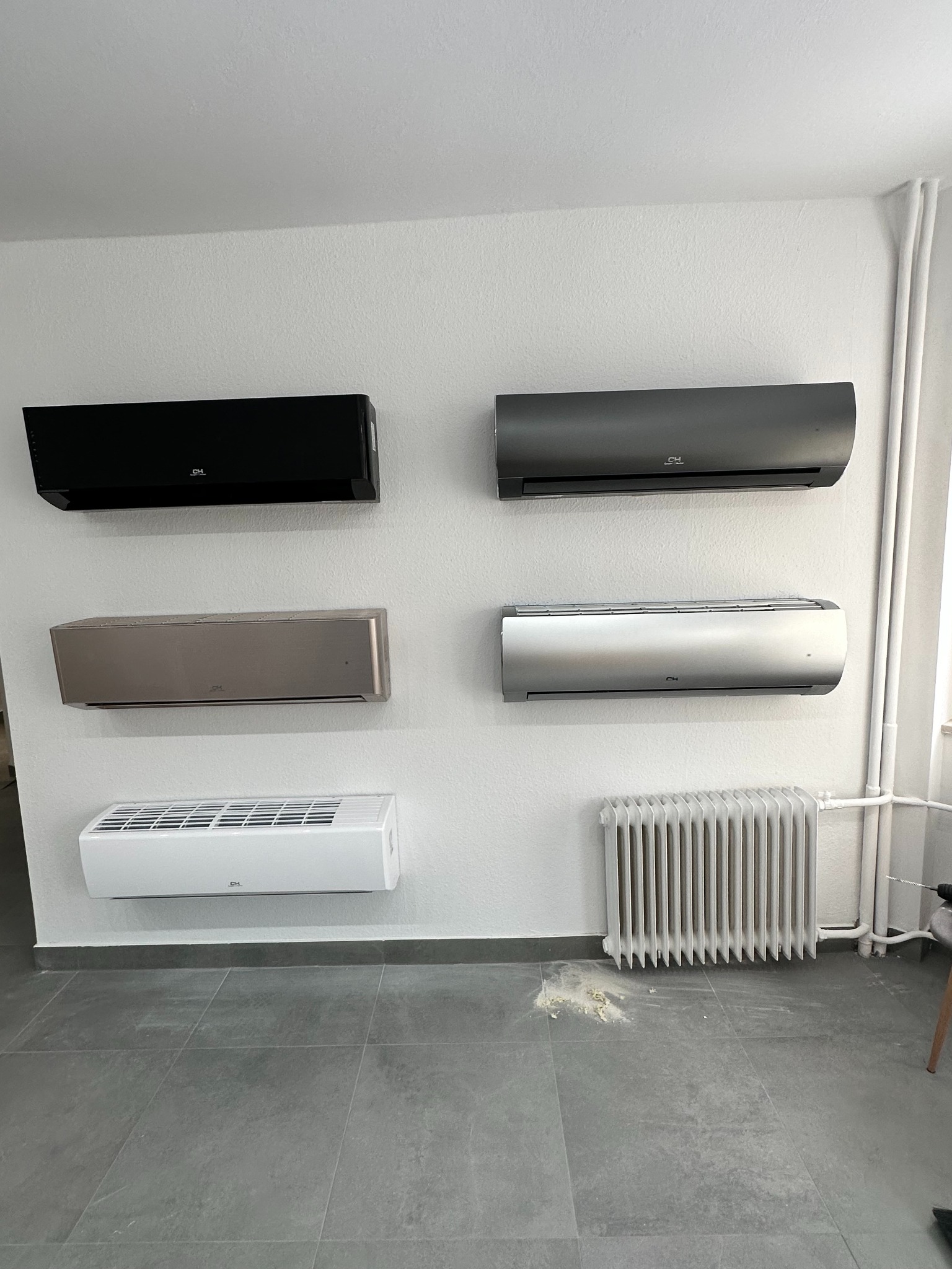 Prezentacja różnych modeli klimatyzatorów ściennych marki CH w kolorach czarnym, szarym, srebrnym, złotym i białym, zainstalowanych na białej ścianie w pomieszczeniu z szarą podłogą i grzejnikiem.