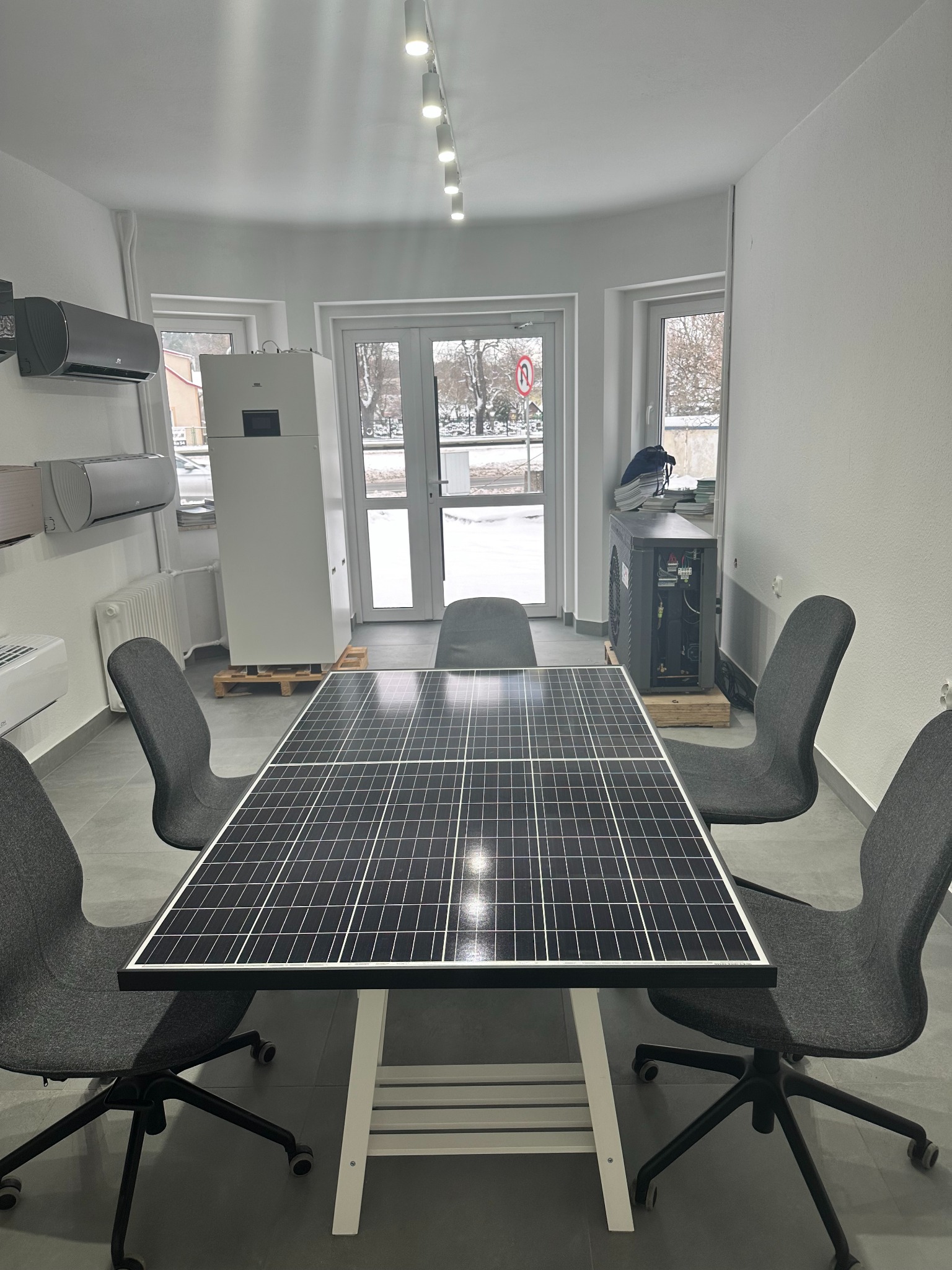Wnętrze biura z panelem fotowoltaicznym umieszczonym na stole otoczonym krzesłami, widoczna pompa ciepła i klimatyzatory na ścianie, za oknem zimowy krajobraz.