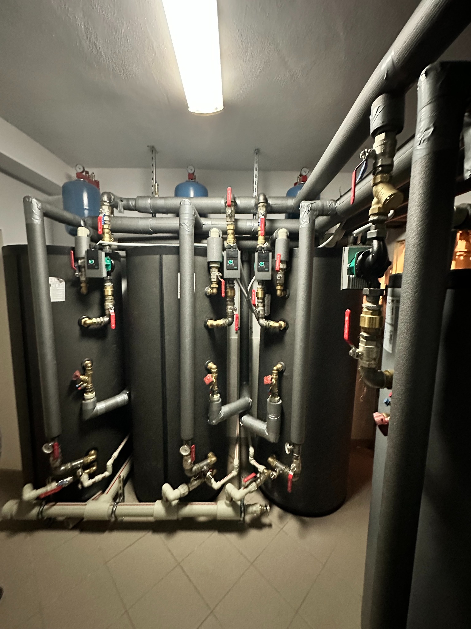 Kotłownia z trzema czarnymi zbiornikami akumulacyjnymi ciepła, licznymi rurami izolowanymi szarą pianką, zaworami z czerwonymi i niebieskimi oznaczeniami, oraz elementami sterującymi z zielonymi...