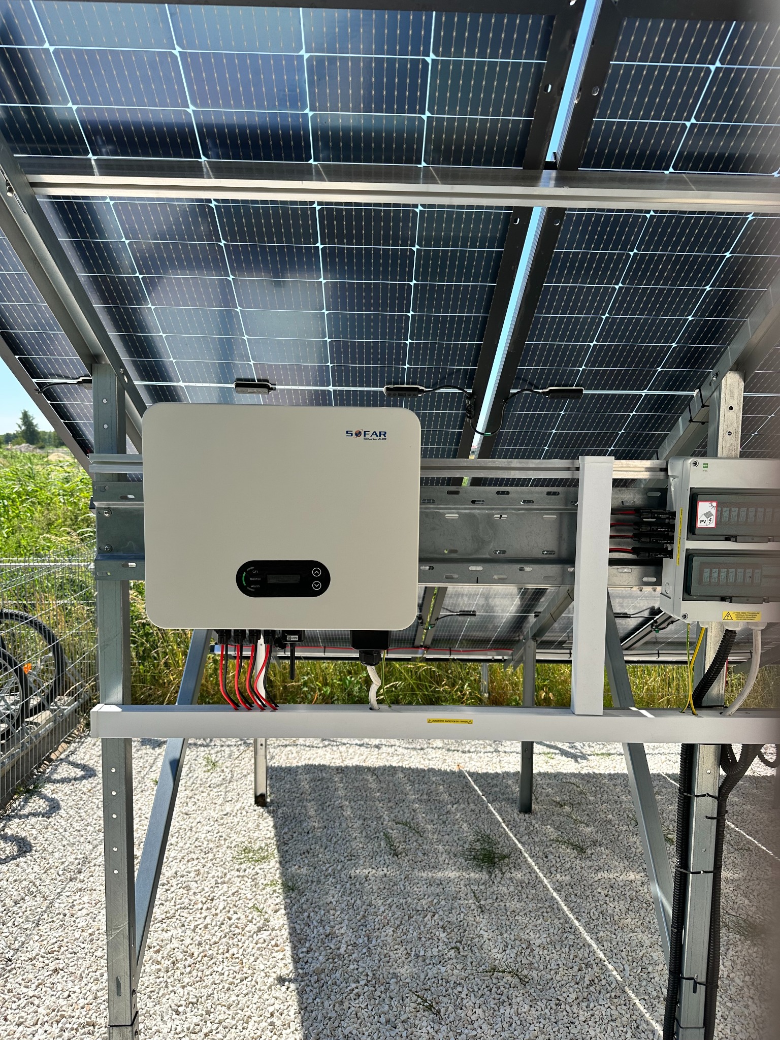 Montaż paneli fotowoltaicznych z widocznym inwerterem SOFAR na metalowej konstrukcji, okablowanie, żwirowe podłoże i zieleń w tle.