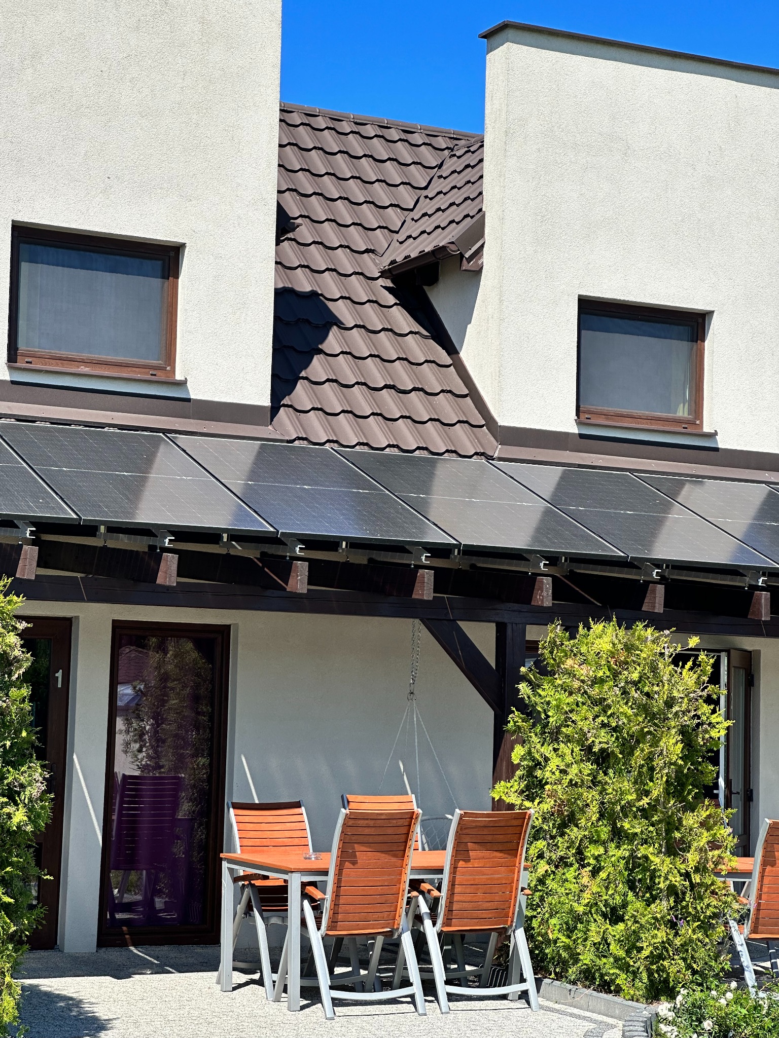 Dom z panelami słonecznymi nad tarasem, brązowy dach, jasna elewacja, drewniane meble ogrodowe i bujna zieleń.