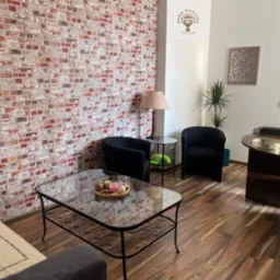 Gabinet z ceglaną ścianą, kanapą, szklanym stolikiem kawowym z dekoracjami, dwoma czarnymi fotelami i biurkiem z roślinami.