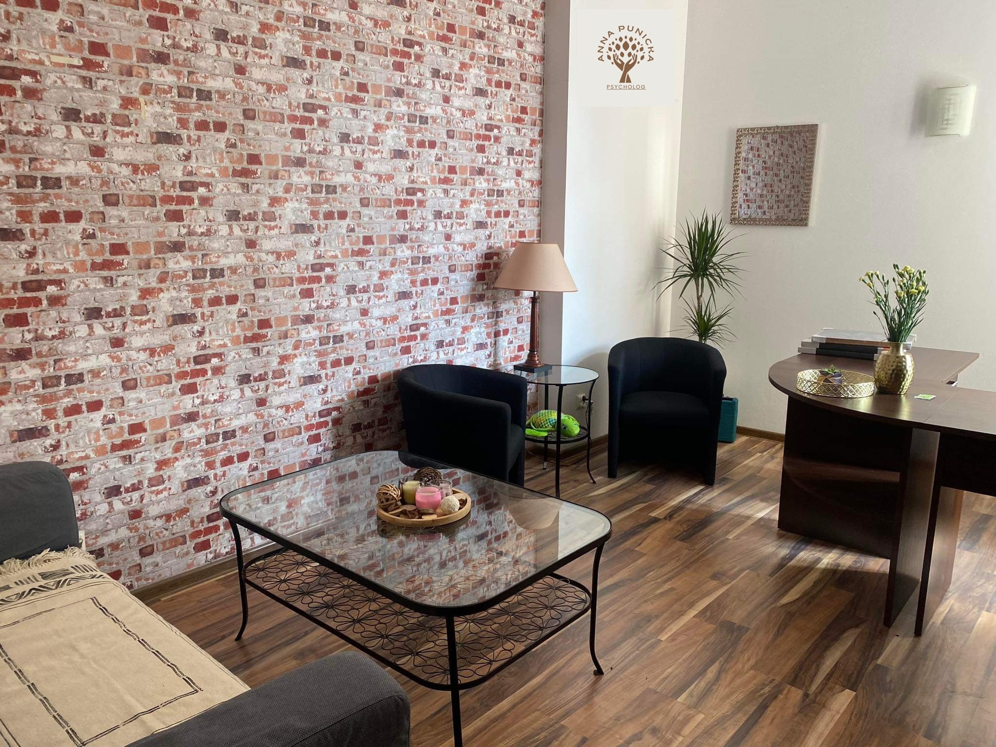 Gabinet z ceglaną ścianą, kanapą, szklanym stolikiem kawowym z dekoracjami, dwoma czarnymi fotelami i biurkiem z roślinami.