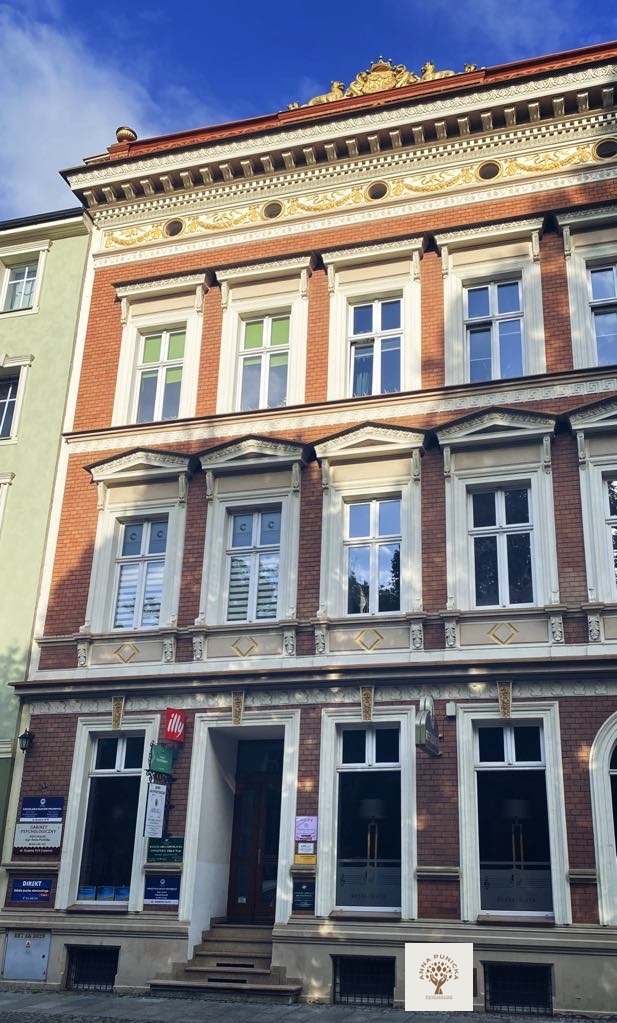 Fasada kamienicy z czerwonej cegły z oknami i zdobieniami, na parterze wejście do lokalu z szyldami i logo firmy.