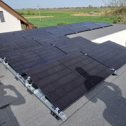 Instal Damian Kostrzewa - Panele słoneczne zamontowane na płaskim dachu budynku, widok z góry, z widocznym kominem z czerwonej cegły i okolicznym krajobrazem.