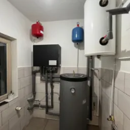 Zestaw instalacji grzewczej z szarym zbiornikiem akumulacyjnym, czarnym modułem sterującym, białym zasobnikiem ciepłej wody i dwoma zbiornikami wyrównawczymi w kolorach czerwonym i niebieskim...