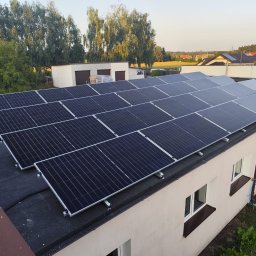 Instal Damian Kostrzewa - Rząd paneli słonecznych na płaskim dachu budynku z oknami, widok z góry, w tle drzewa i budynki mieszkalne.