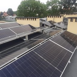 Instal Damian Kostrzewa - Instalacja paneli słonecznych na płaskim dachu budynku z żółtymi kominami wentylacyjnymi; widok z góry, w tle zabudowa miejska.