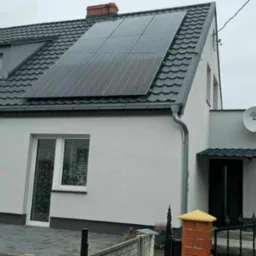 Dom jednorodzinny z szarą dachówką i panelami słonecznymi na dachu, widziany zza ogrodzenia z ceglanym słupkiem.