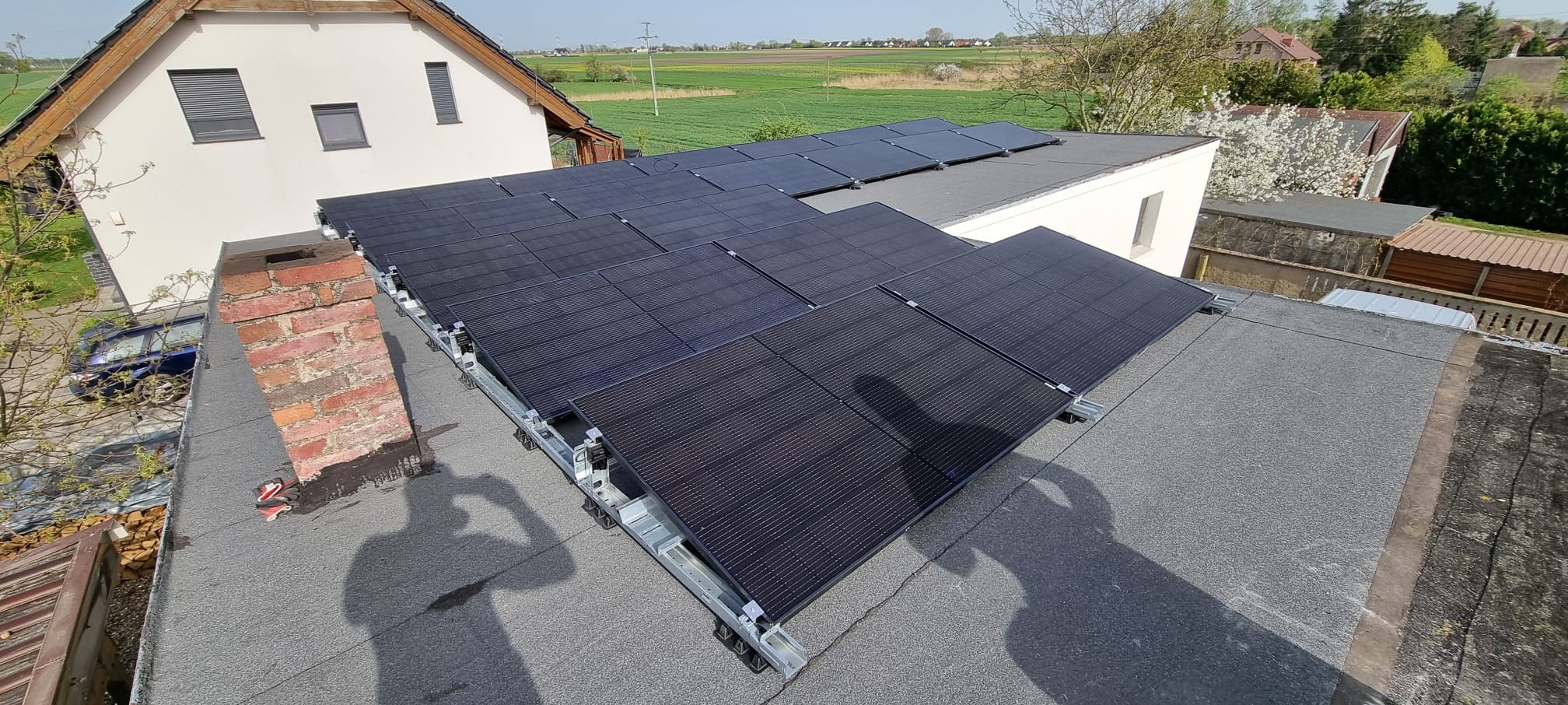 Panele słoneczne zamontowane na płaskim dachu budynku, widok z góry, z widocznym kominem z czerwonej cegły i okolicznym krajobrazem.