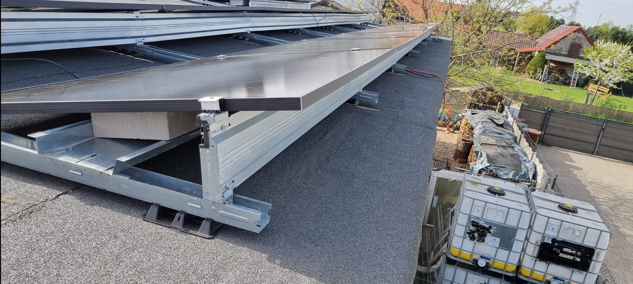 Montaż paneli słonecznych na dachu z widocznymi elementami konstrukcyjnymi i obciążeniem z bloczków betonowych, w tle zabudowania gospodarcze i ogród.