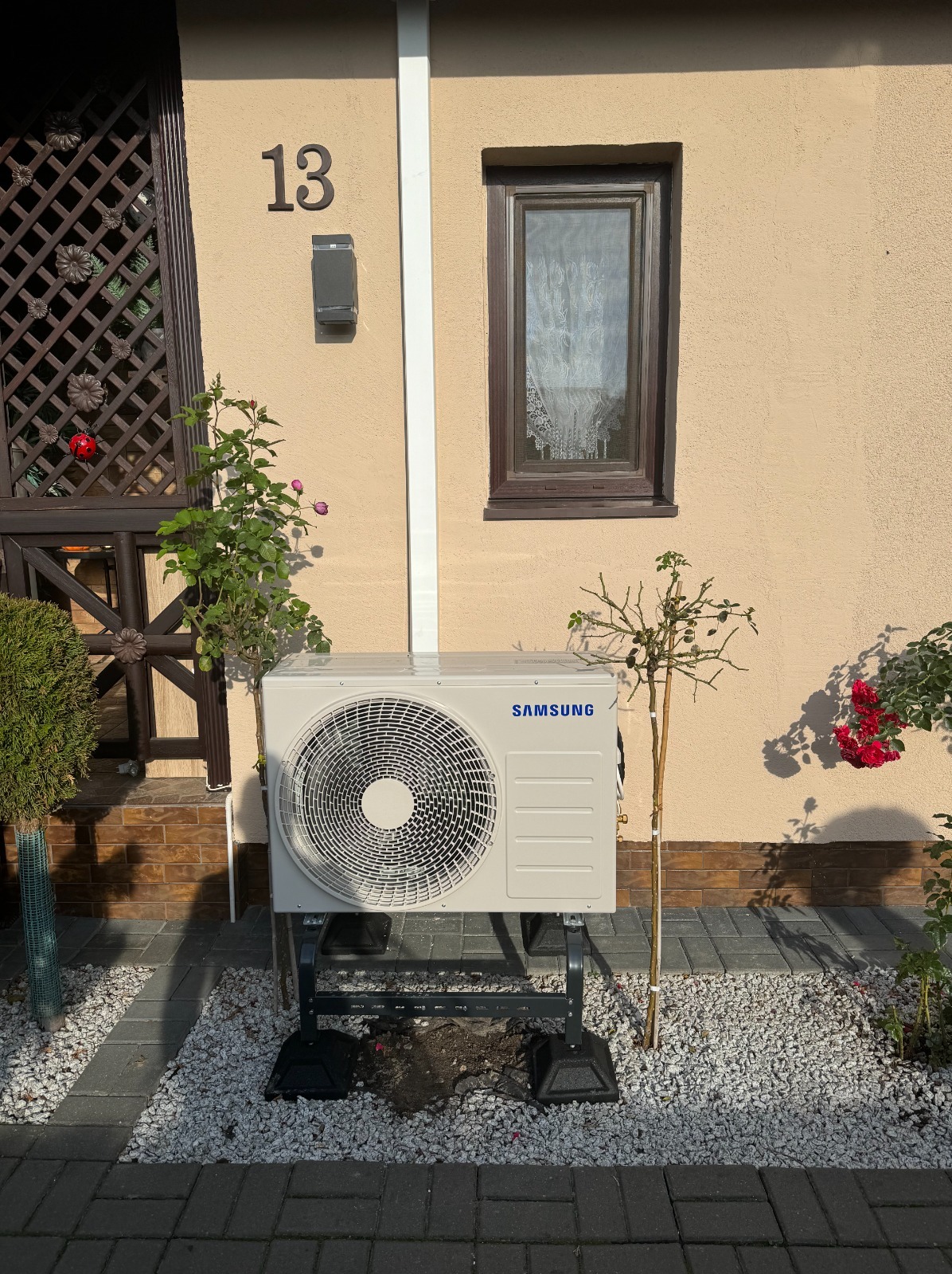 Zewnętrzna jednostka pompy ciepła Samsung zamontowana na metalowej konstrukcji przed beżową ścianą domu z numerem 13, otoczona roślinami i białym żwirem.