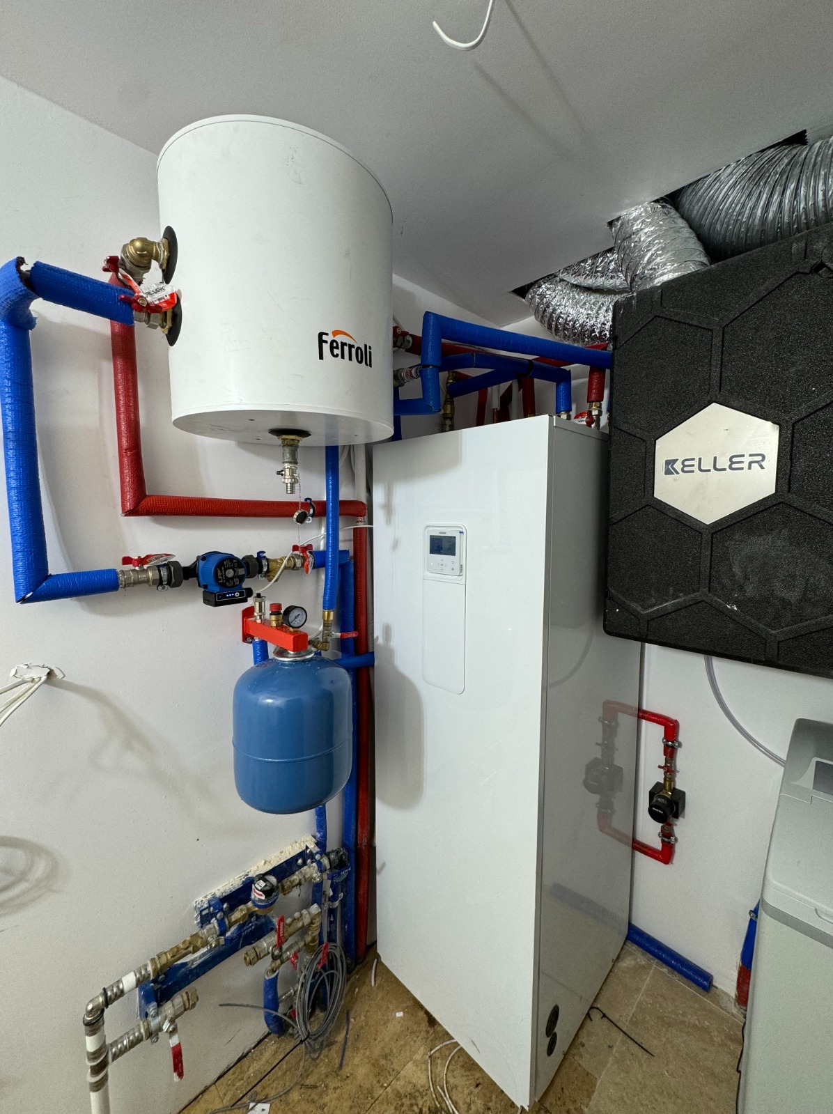 Instalacja pompy ciepła w kotłowni z widocznym zbiornikiem buforowym Ferroli, modułem Keller i licznymi rurami w izolacji niebieskiej i czerwonej.