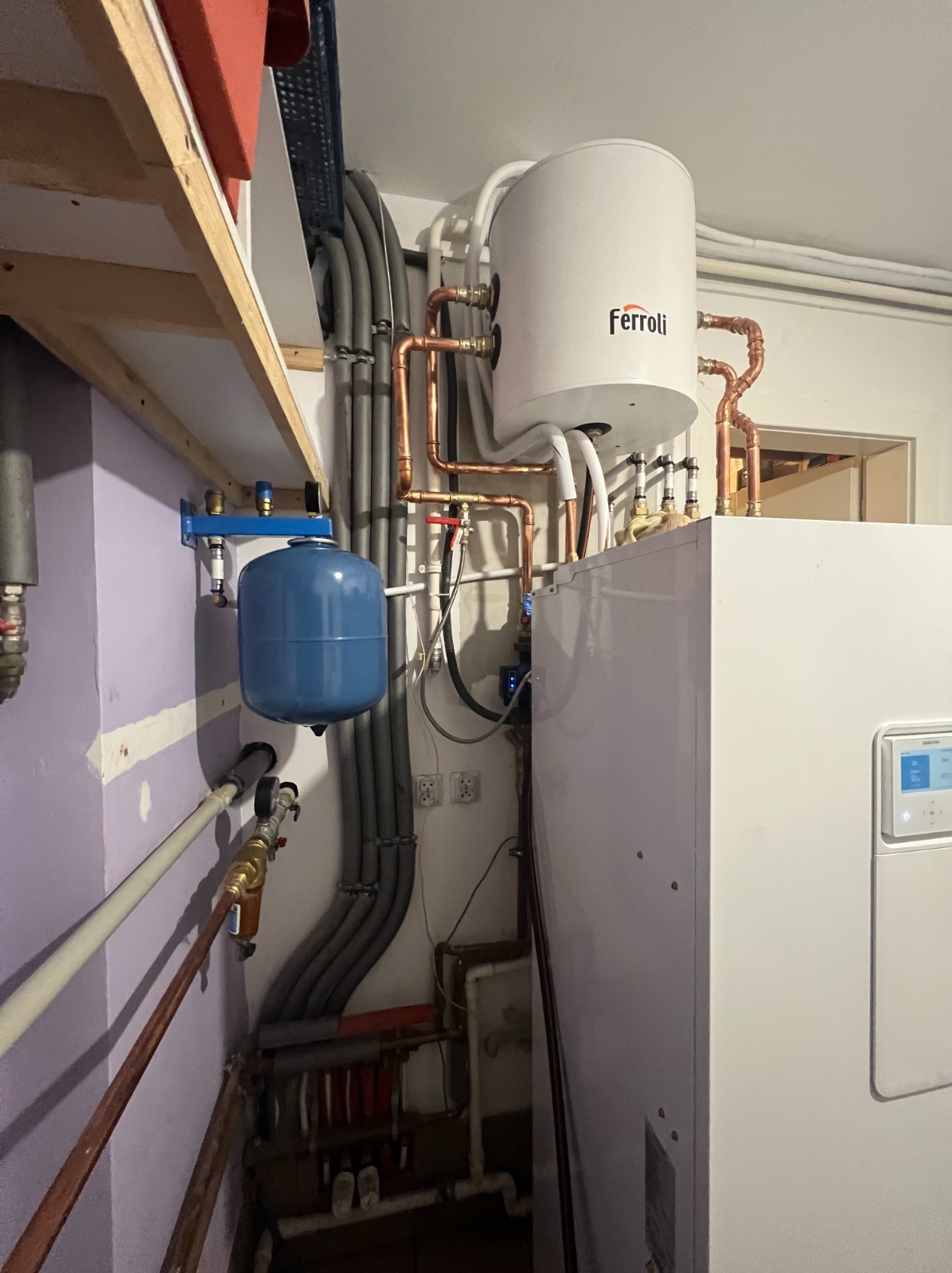 Instalacja grzewcza w kotłowni: biały zasobnik Ferroli, niebieskie naczynie przeponowe, miedziane rury i szare przewody, widoczne gniazdka elektryczne na ścianie.