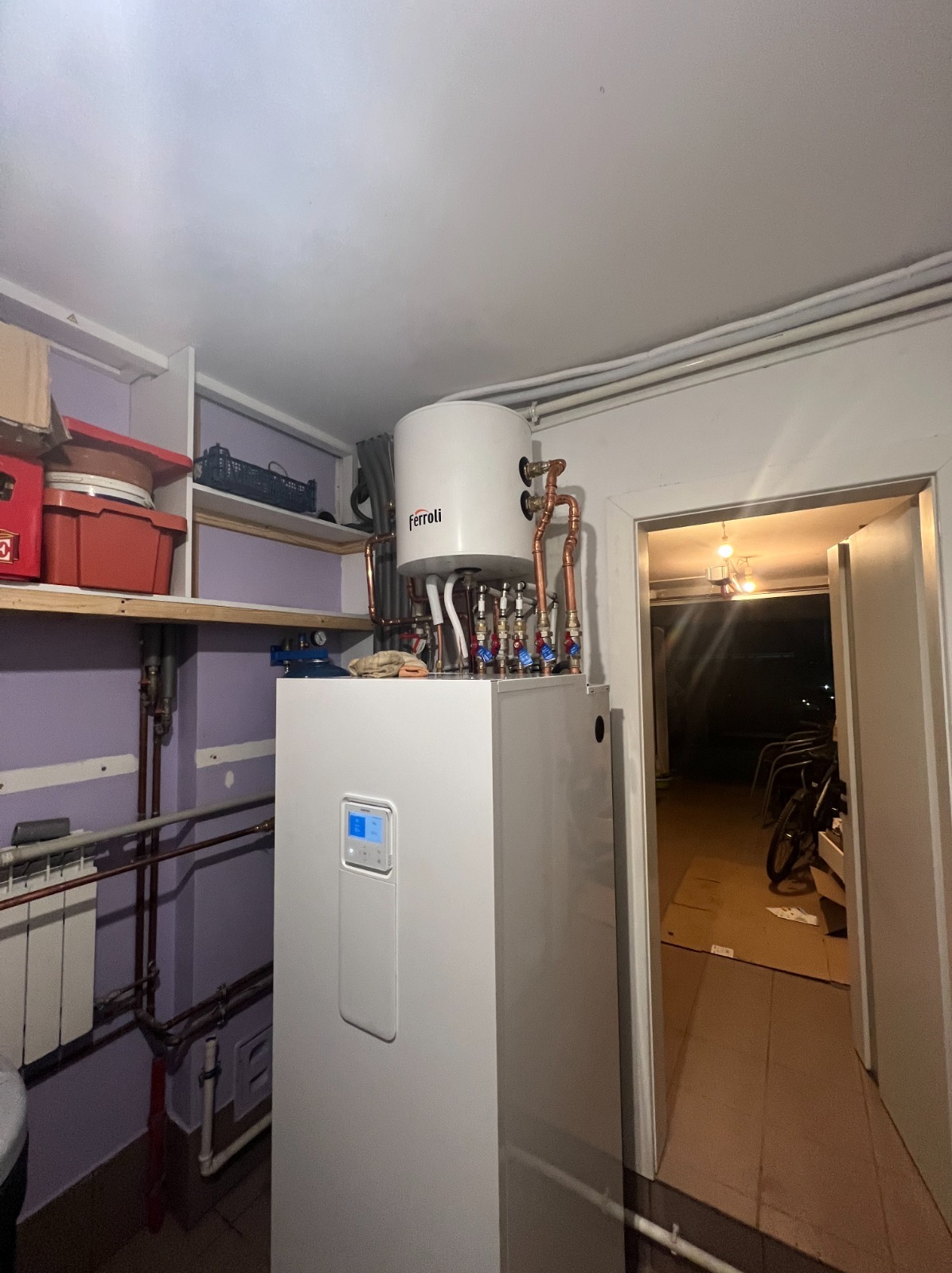 Biała pompa ciepła Ferroli z widocznymi miedzianymi rurami i zaworami, zainstalowana w pomieszczeniu z półkami i widokiem na garaż.