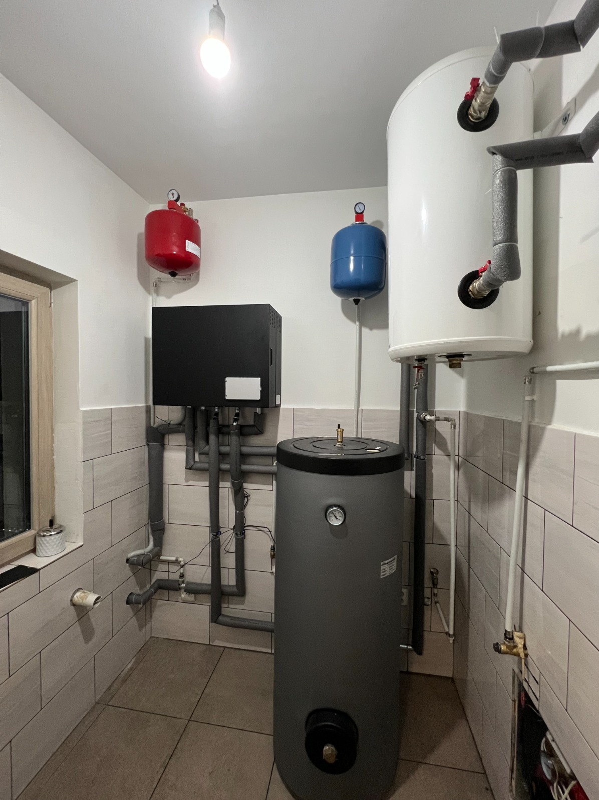 Zestaw instalacji grzewczej z szarym zbiornikiem akumulacyjnym, czarnym modułem sterującym, białym zasobnikiem ciepłej wody i dwoma zbiornikami wyrównawczymi w kolorach czerwonym i niebieskim...