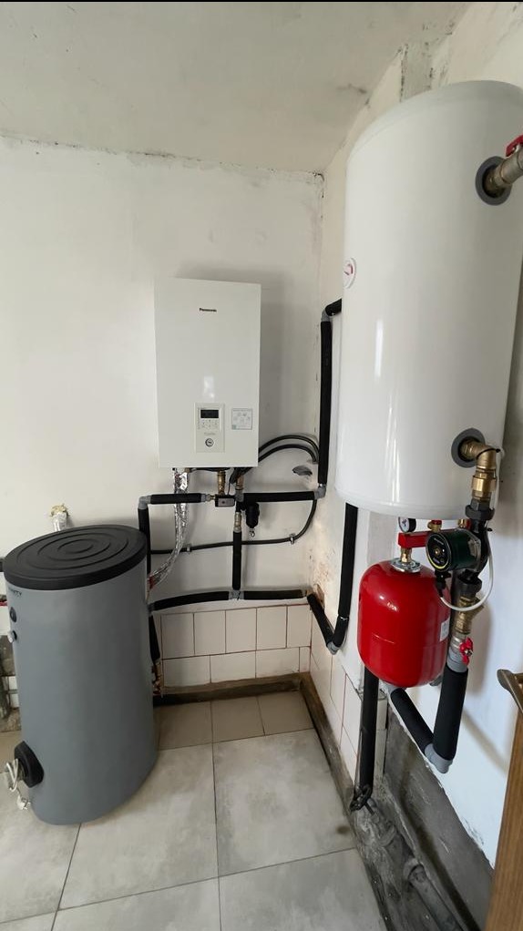 Biały kondensacyjny kocioł gazowy Panasonic, biały zasobnik ciepłej wody i szary zbiornik buforowy połączone czarnymi rurami w pomieszczeniu z białymi ścianami i beżowymi płytkami podłogowymi.