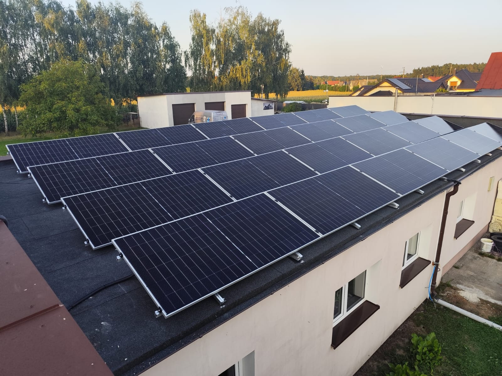 Rząd paneli słonecznych na płaskim dachu budynku z oknami, widok z góry, w tle drzewa i budynki mieszkalne.