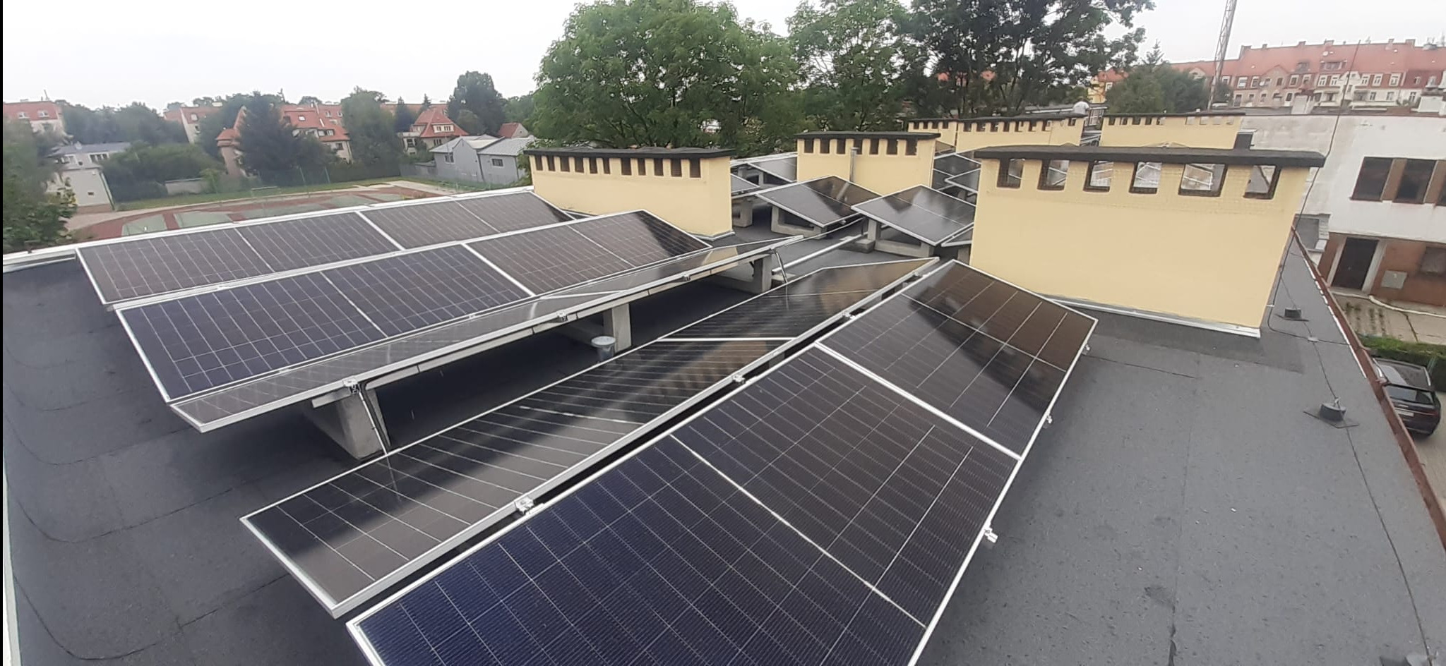 Instalacja paneli słonecznych na płaskim dachu budynku z żółtymi kominami wentylacyjnymi; widok z góry, w tle zabudowa miejska.