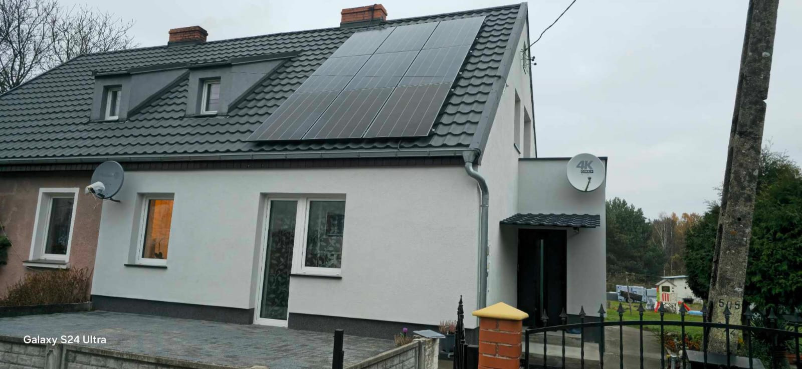 Dom jednorodzinny z szarą dachówką i panelami słonecznymi na dachu, widziany zza ogrodzenia z ceglanym słupkiem.