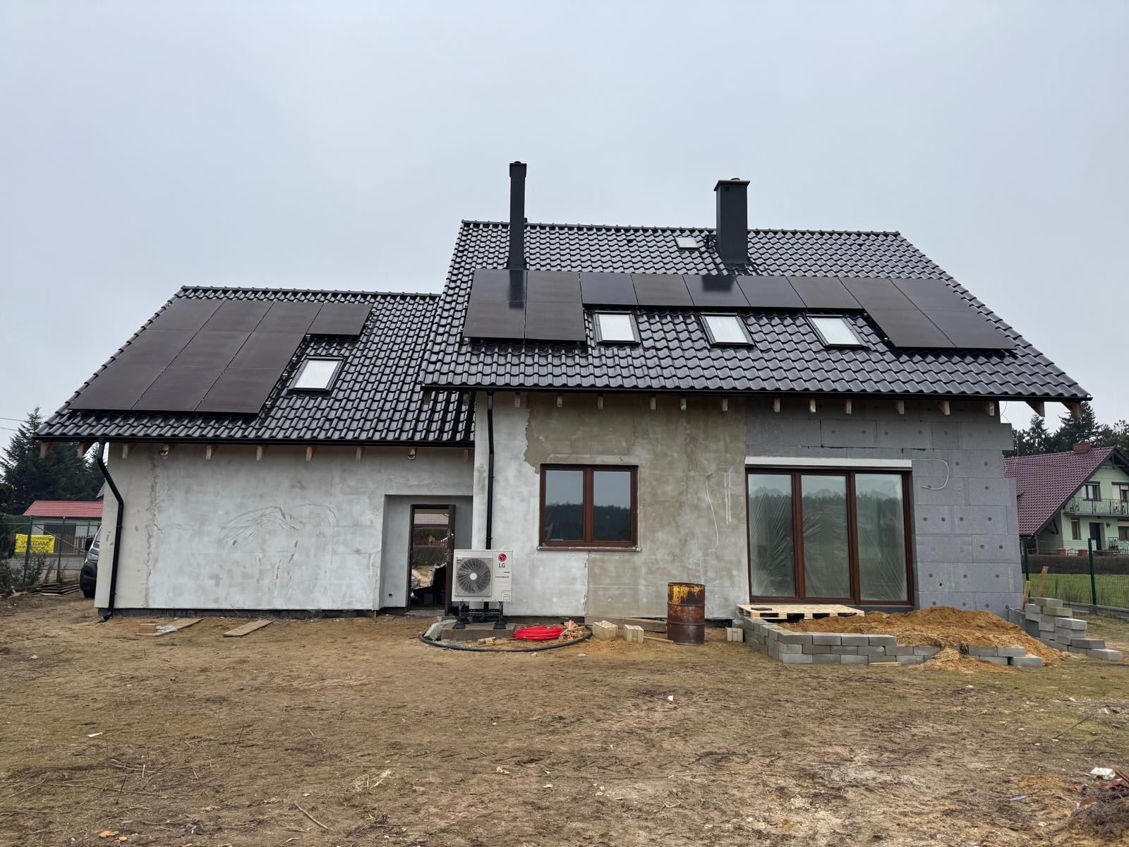 Dom jednorodzinny w trakcie wykańczania z zamontowanymi panelami słonecznymi na dachu i pompą ciepła LG na zewnątrz, widoczne okna dachowe, elewacja w stanie surowym.