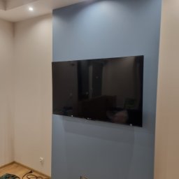 Ruslan - Ściana pomalowana na dwa kolory z zamontowanym telewizorem, widoczne gniazdka do podłączenia sprzętu, w tle widać narzędzia i kabel zasilający