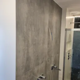 Szara ściana z płytek imitujących beton, z wystającymi przewodami elektrycznymi i zamontowaną armaturą łazienkową w nowoczesnej łazience z kabiną prysznicową.