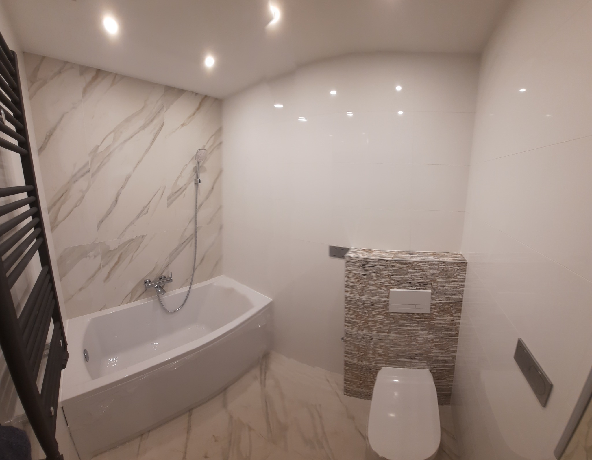 Nowoczesna łazienka z wanną, grzejnikiem drabinkowym i podwieszaną toaletą, wykończona płytkami imitującymi marmur i mozaiką.