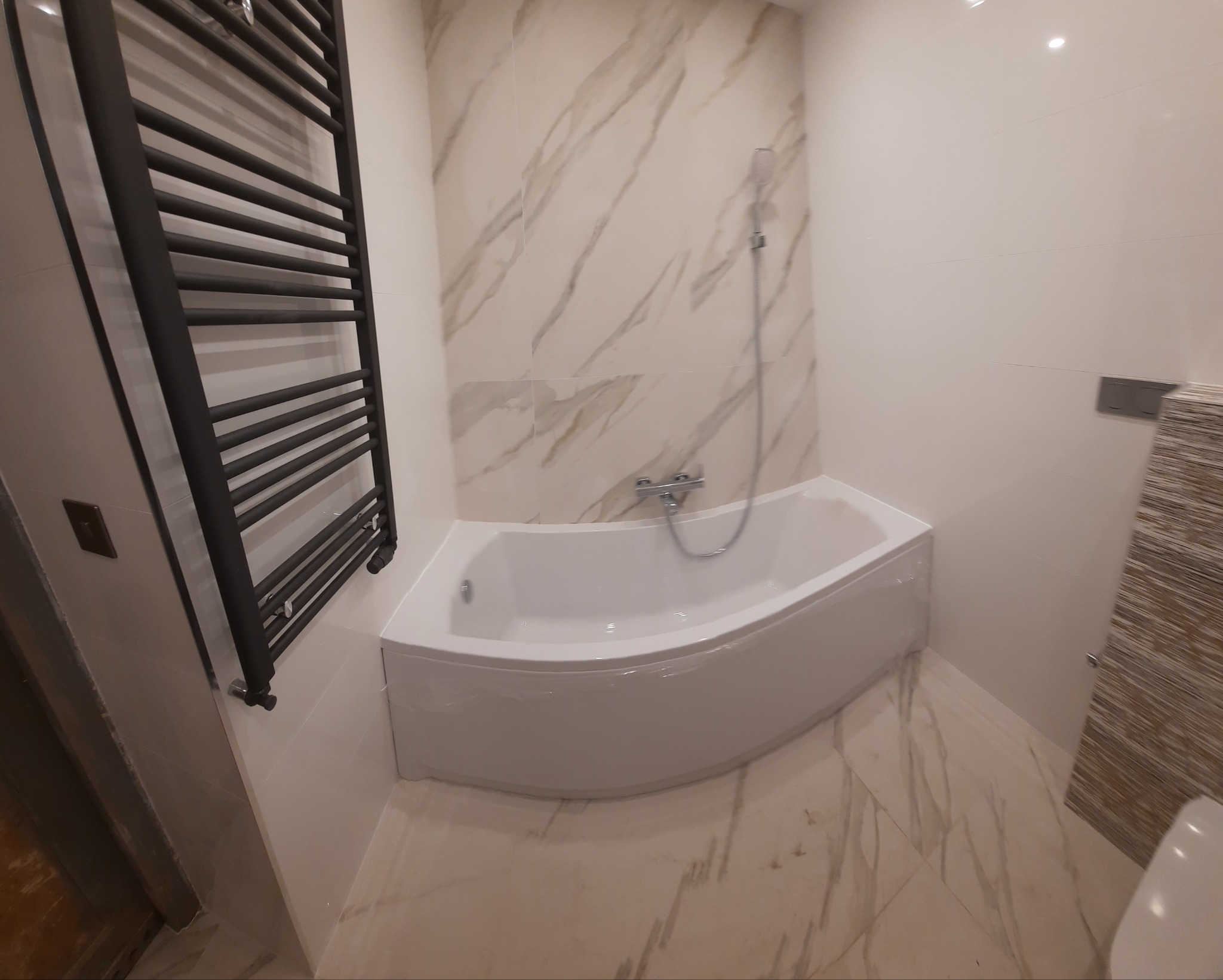 Nowoczesna łazienka z narożną wanną obudowaną białą płytą, marmurowe płytki na ścianach i podłodze, czarny grzejnik drabinkowy po lewej stronie.