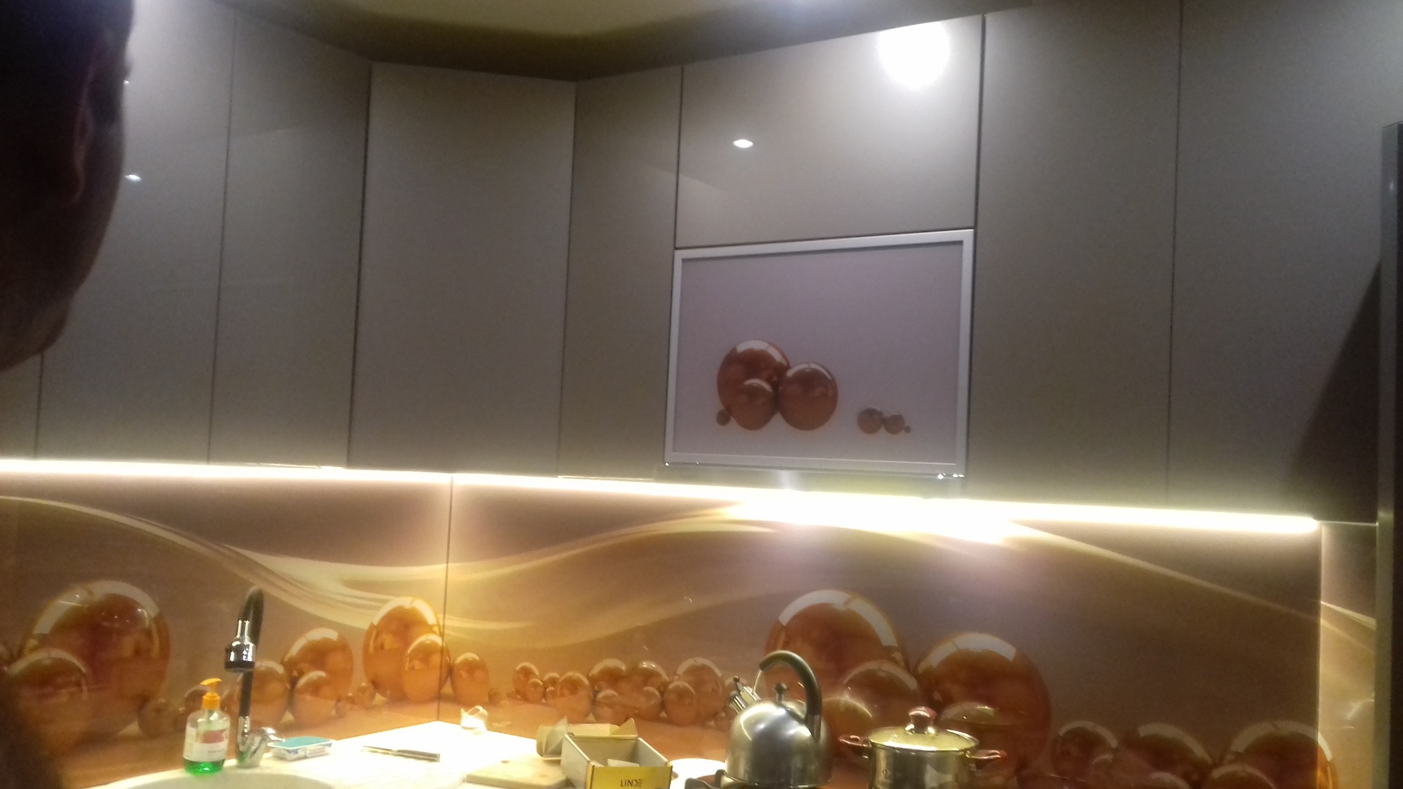 Nowoczesna kuchnia z szarymi szafkami, podświetleniem LED i panelem szklanym z motywem kul, widok na zlewozmywak, baterię i czajnik.