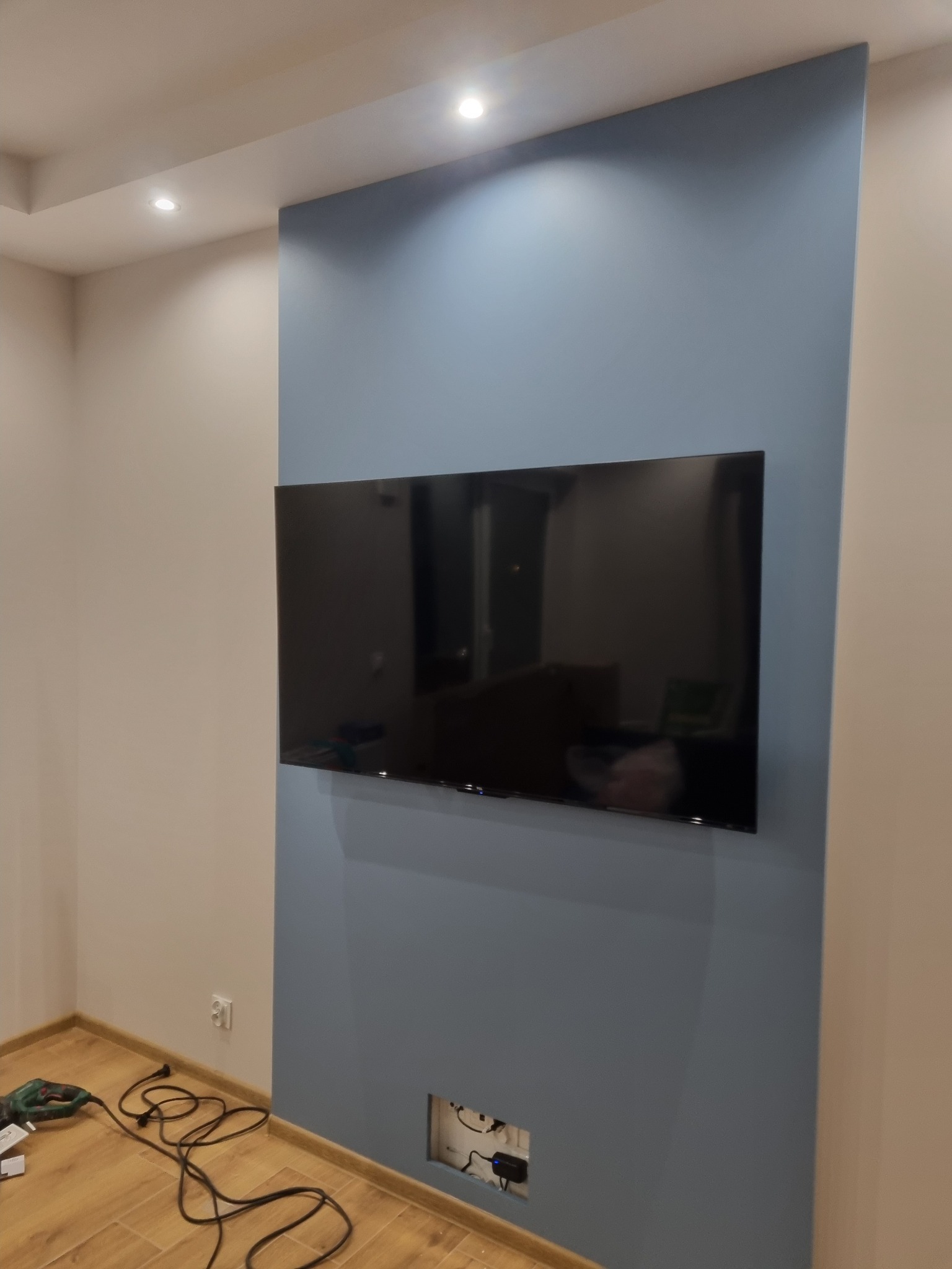 Ściana pomalowana na dwa kolory z zamontowanym telewizorem, widoczne gniazdka do podłączenia sprzętu, w tle widać narzędzia i kabel zasilający