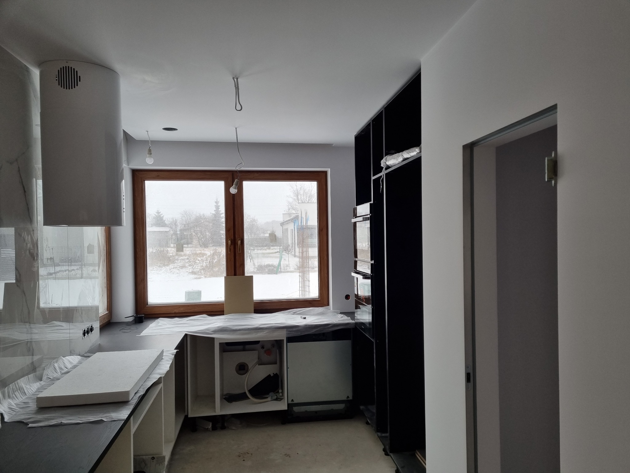 Wnętrze kuchni w trakcie montażu: biały okap cylindryczny, czarne szafki, okno z widokiem na zimowy krajobraz, elementy zabezpieczone folią.