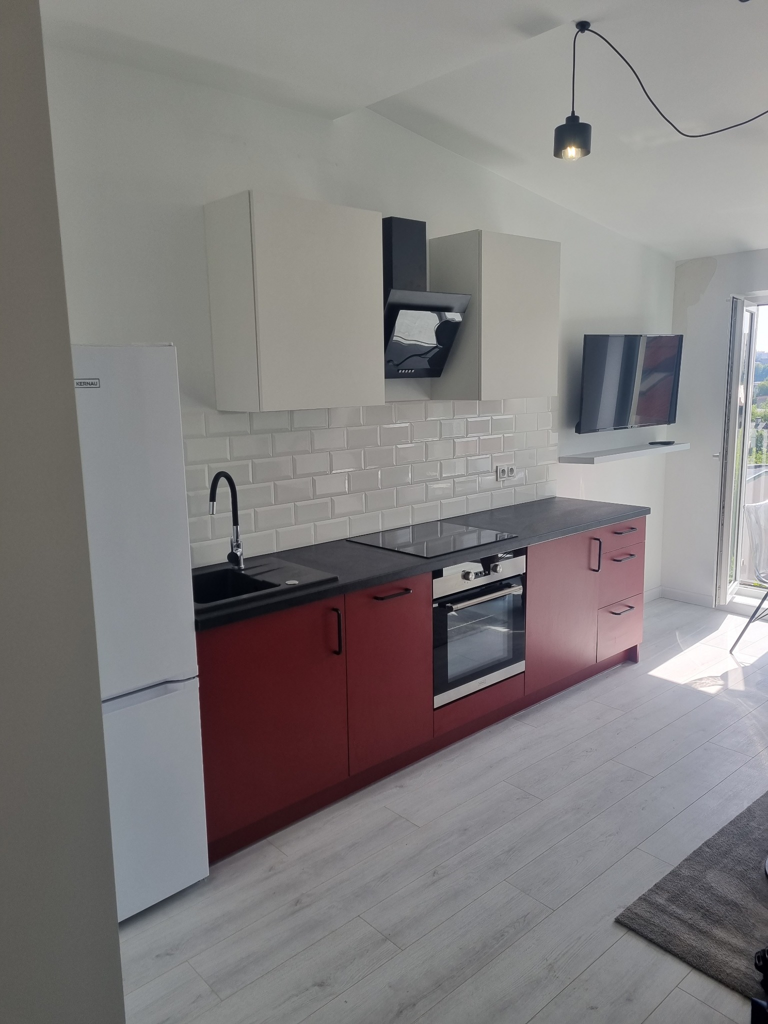 Nowoczesna kuchnia z białymi szafkami górnymi i bordowymi dolnymi, białymi płytkami metra na ścianie i czarnym blatem, widok na balkon.