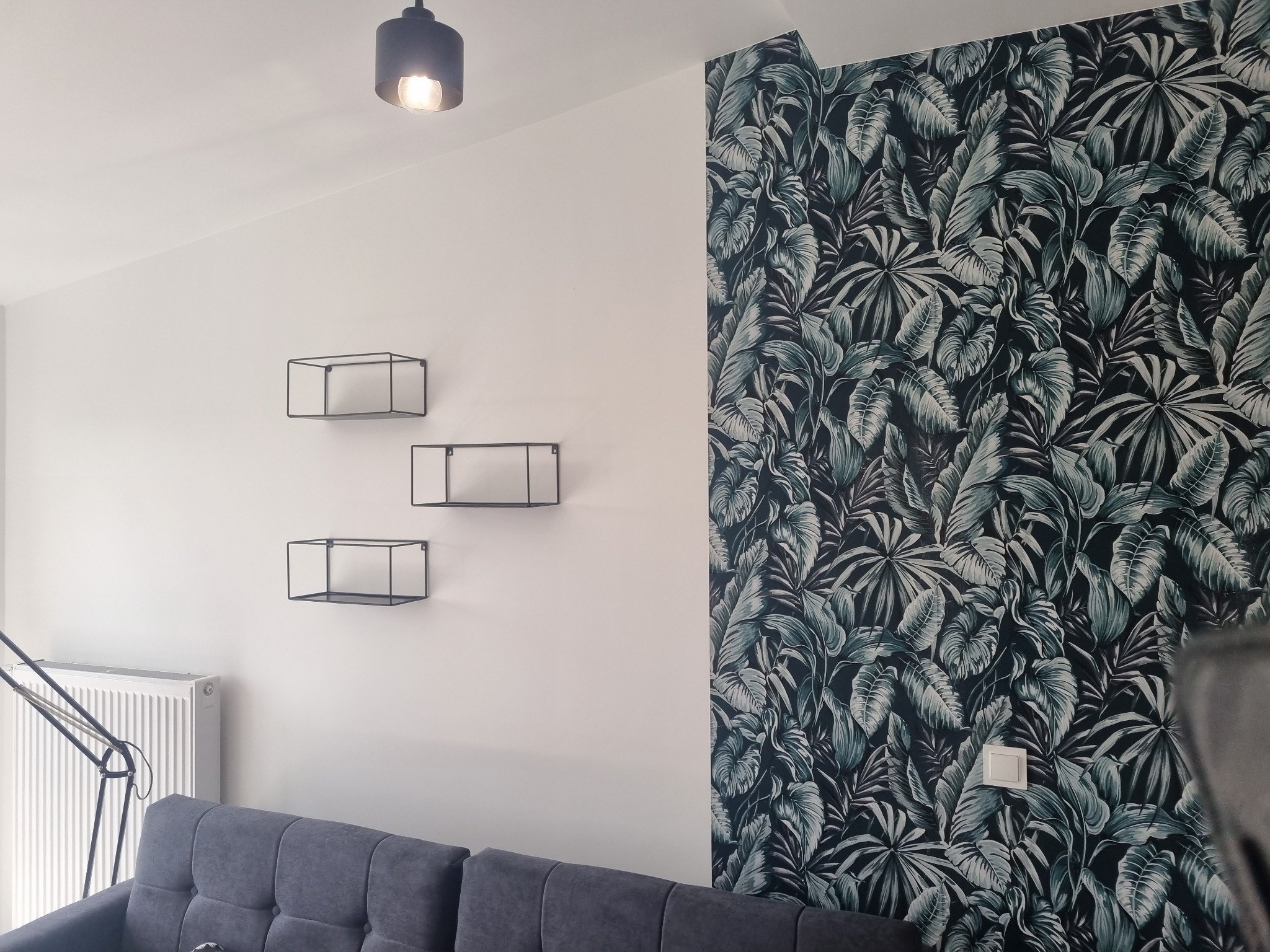 Pokój z fragmentem ściany pokrytym tapetą w tropikalne liście, trzema metalowymi półkami ściennymi w kształcie prostopadłościanów, szarą sofą, lampą podłogową i grzejnikiem.